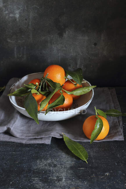 Mandarini con foglie in ciotola e su tavola con panno — Foto stock