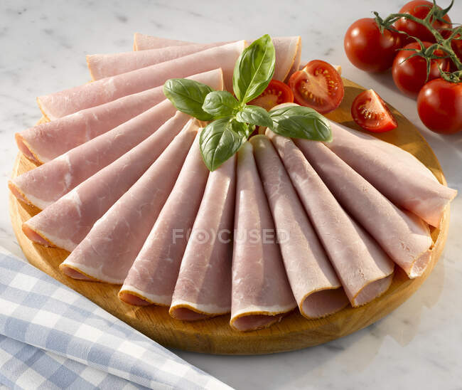 Slices of honey roasted ham on a circular wooden cutting board with garnish of basil and tomatoes — Stock Photo