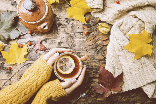 Чашка горячего чая с лимоном в руках женщины, держа его на деревянном осеннем фоне — стоковое фото