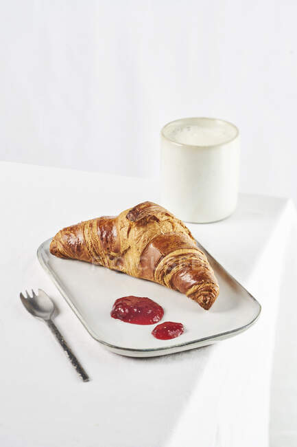 Croissant with cherry jam on plate with fork and cup of milk on table — Stock Photo