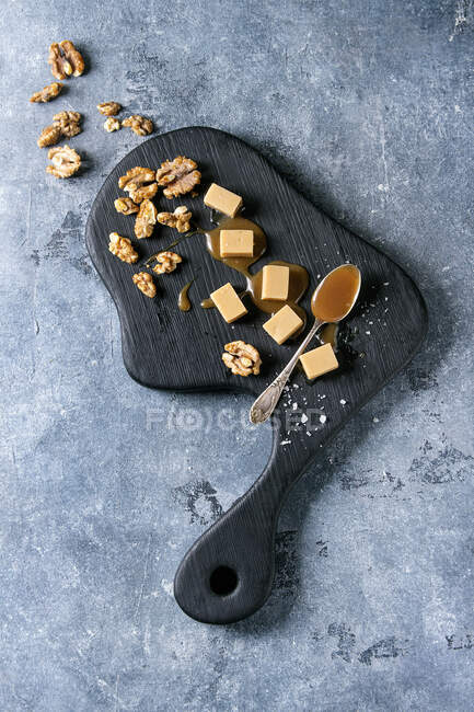 Salted caramel fudge candy served on black wooden board with fleur de sel, caramel sauce and caramelized walnuts — Stock Photo