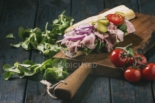 Beef and vegetables sandwich with sliced meat, pickled cucumber, green salad on wooden cutting board over dark kitchen table — Stock Photo