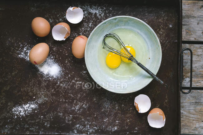 Eggs and eggs shells on baking tray, bowl with liquid eggs and whisk — Stock Photo