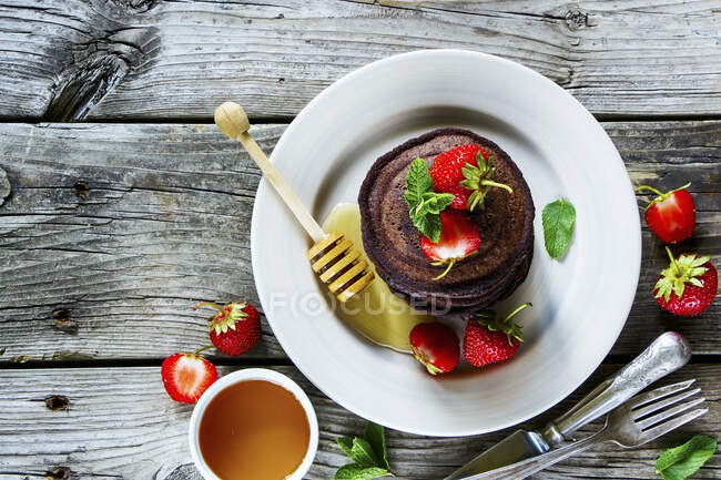 Chocolate pancakes with fresh strawberries and honey, served on ceramic plate over old grey wooden background — Stock Photo