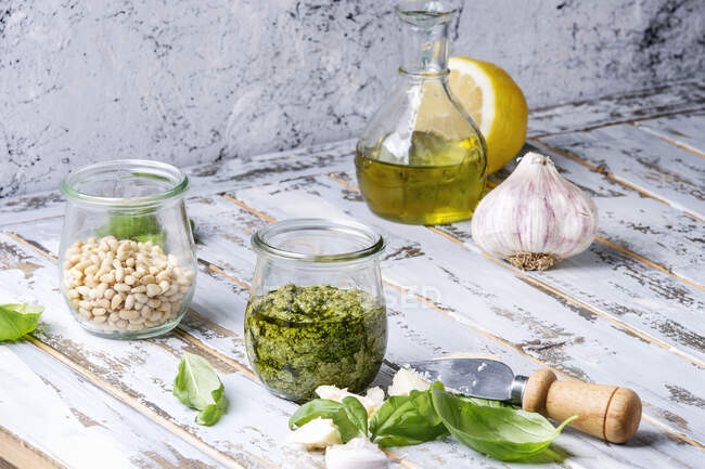 Basil pesto sauce in glass jar with fresh basil, olive oil, parmesan cheese, garlic, pine nuts, lemon on wooden kitchen table — Stock Photo