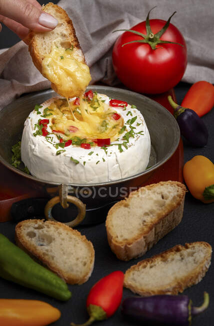 Woman hand dipping slice of bread inside melted Camembert cheese, seasoned with herbs and peppers — Stock Photo