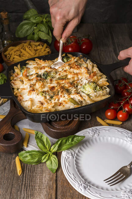 Gebackene Nudeln mit Brokkoli, Blumenkohl, Käse und Bechamelsauce in einer Pfanne mit menschlichen Händen im Rahmen auf Holzgrund — Stockfoto