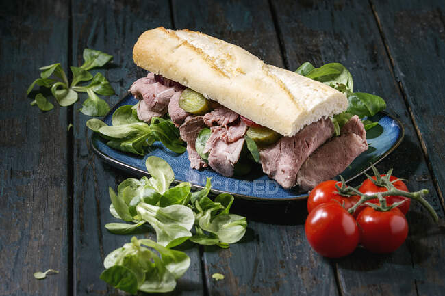 Sándwich de carne de res y verduras con carne en rodajas, pepino en escabeche, ensalada verde en plato de cerámica azul - foto de stock