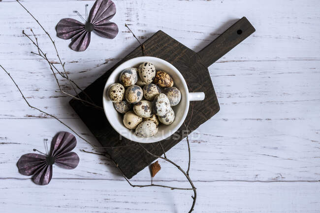 Top view of quail eggs in a white bowl on a black cutting board decorated with a bare tree branch and paper butterflies — Stock Photo