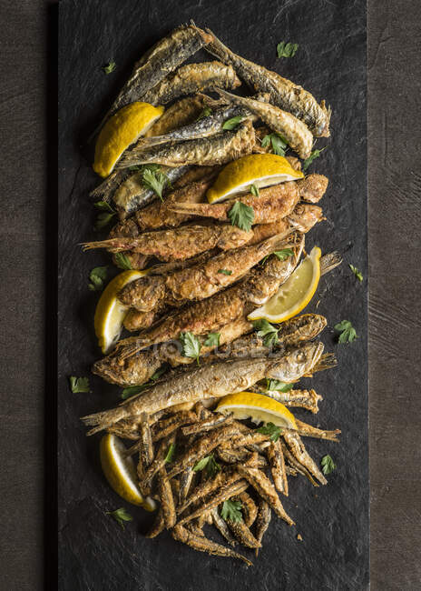 Ассортимент жареной рыбы с лимонными клинками и петрушкой на доске и темном фоне — стоковое фото