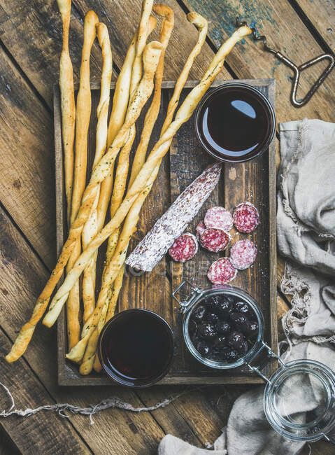 Italian Grissini bread sticks, dry cured pork meat sausage, black olives in jar and red wine in glasses - foto de stock