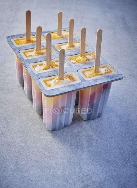 Tri-coloured ice cream sticks in lolly moulds on a grey surface — Stock Photo