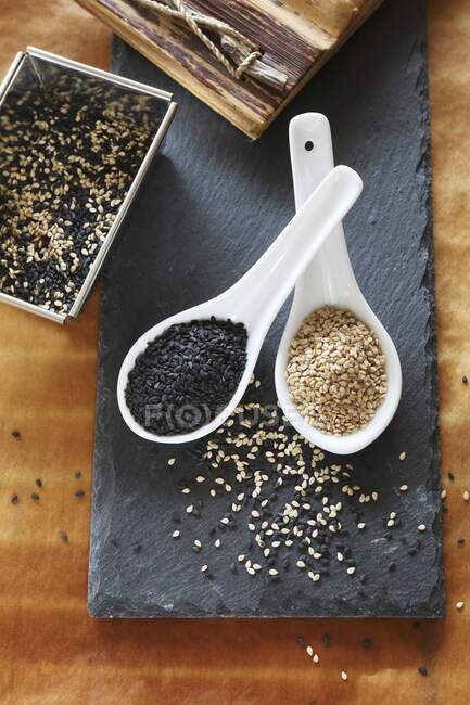 Семена черного и белого кунжута в ложках на тарелке с шифер — стоковое фото