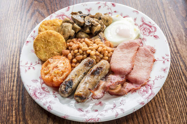 Традиционный английский завтрак с беконом, сосисками, помидорами и грибами, запечёнными бобами, картофельными оладьями и яичницей на деревянном столе — стоковое фото