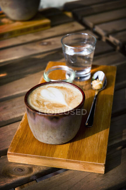 Um cappucino em um copo de barro rústico em uma placa de madeira com um copo de água e açúcar — Fotografia de Stock