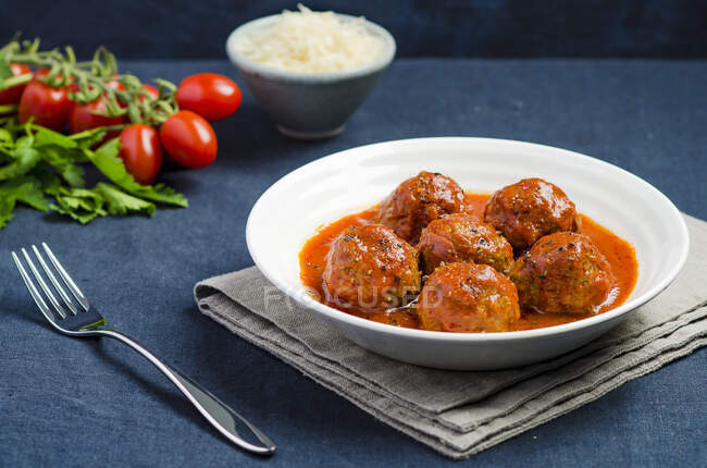 Plate of meatballs in tomato sauce with black pepper on a blue tablecloth with fork and ingredients in the background — Stock Photo