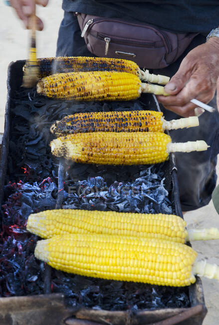 Um vendedor de rua balinês que vende espigas de milho picantes em um churrasco a carvão com um cigarro na mão — Fotografia de Stock