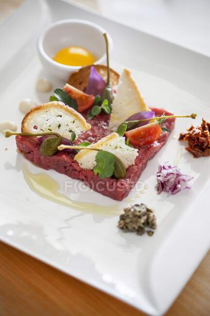 Beef tartare with capers, chopped onion, dried tomatoes in oil and croutons, next to egg yolk in bowl — Stock Photo