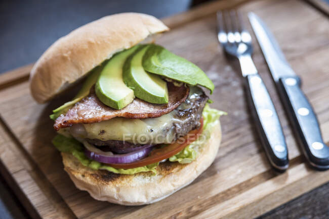Burger with avocado slices, cheese, bacon, onions, lettuce, tomatoes on a wooden board with cutlery — Stock Photo