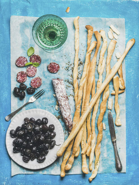 Italian Grissini bread sticks, dry cured pork meat sausage, black olives in white plate and white wine — Stock Photo