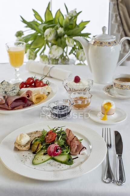 A breakfast table with prosciutto, mixed leaves, balsamic vinegar, bread and butter, cucumber, radishes, a soft boiled egg and a cheese plate — Stock Photo