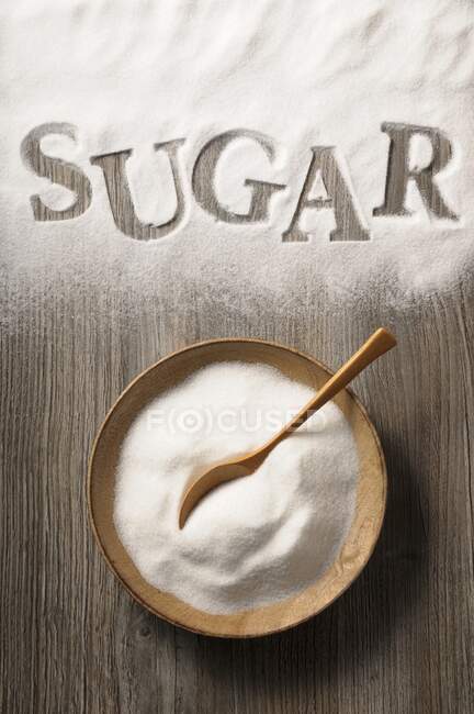 Azúcar en un tazón de madera y derramado sobre un fondo de madera con la palabra 'azúcar' - foto de stock