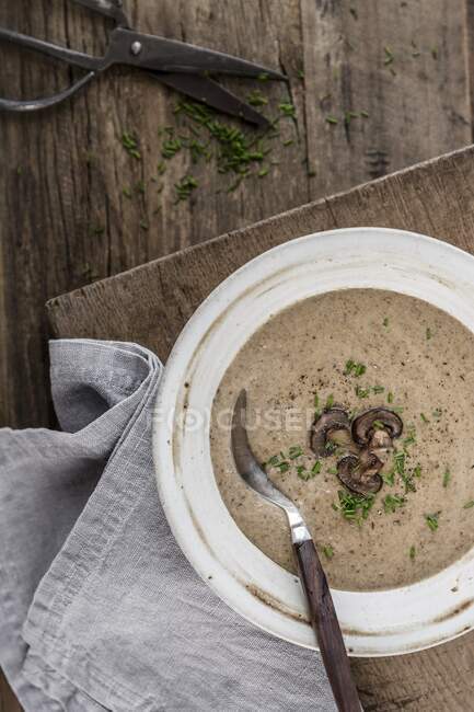 Sopa de champiñones gruesa y cremosa decorada con rodajas de champiñones y cebollino en un tazón blanco sobre una superficie de madera - foto de stock
