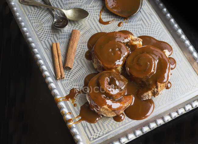 Rollos de canela con cobertura de caramelo caliente y palitos de canela en una bandeja de plata con cucharas de té - foto de stock