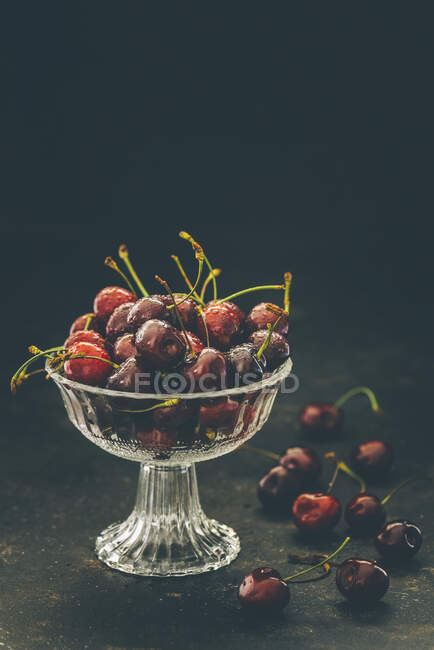 Wet cherries with stems in glass bowl and on dark surface — Stock Photo