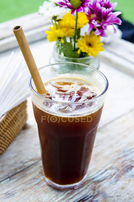An balinese iced coffee with an ecological bamboo straw on a table with flowers in the background — Photo de stock