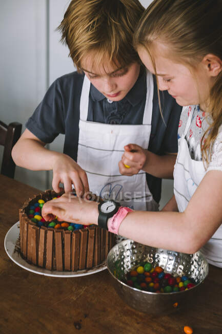 Um menino e uma menina decorando um bolo de chocolate usando aventais brancos — Fotografia de Stock