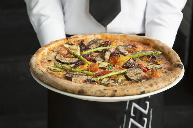 Официант в форме держит овощную пиццу — стоковое фото