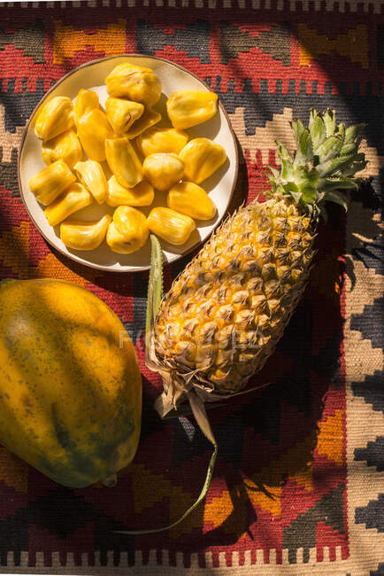 Jackfruchtsegmente auf einem Teller, ganze Ananas und Papaya auf buntem Hintergrund — Stockfoto