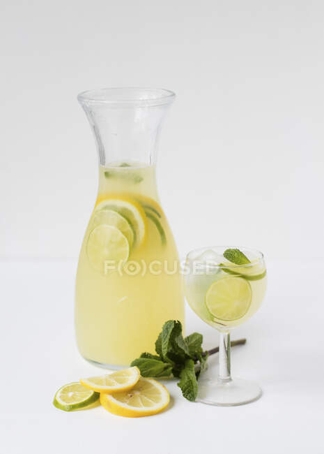 Lemonade in jug and glass with limes, lemons and leaves — Stock Photo