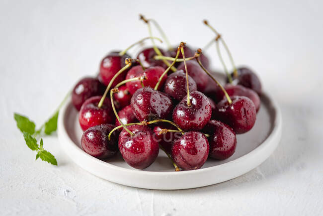Fresh wet cherries on small ceramic plate and mint leaves on table — Stock Photo