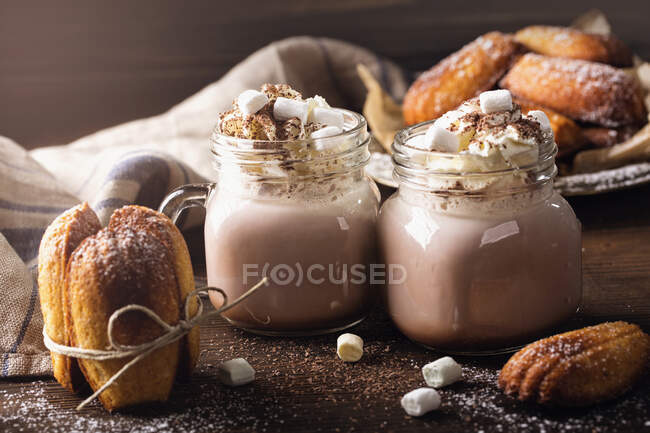 Chocolate caliente con crema batida y malvaviscos servidos con madeleines - foto de stock