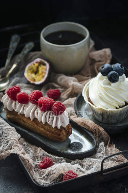 Eclair with berries and cream, and vanilla cupcake, black coffee — Stock Photo