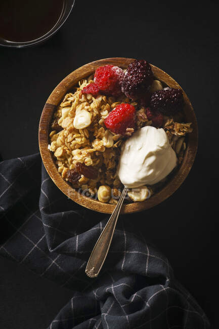 Granola aux framboises et mûres et une cuillère de crème dans un bol en bois avec une tasse de café sur une table noire — Photo de stock