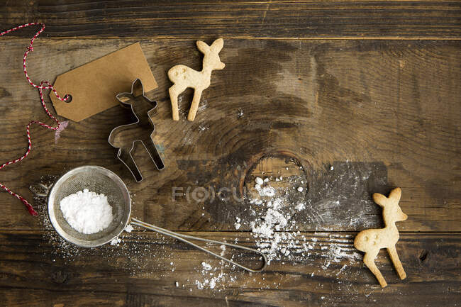 Biscotti natalizi a forma di cervo su tavola rustica con tagliabiscotti, zucchero a velo in setaccio ed etichetta regalo — Foto stock