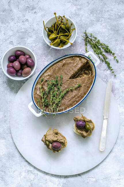 Pâte de foie en céramique, décorée de thym frais et deux sandwichs baguette avec pâté, câpres, olives sur un support en marbre — Photo de stock