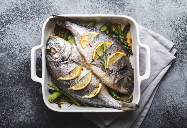 Baked fish dorado with green asparagus in white ceramic baking pan on gray rustic concrete background — Stock Photo