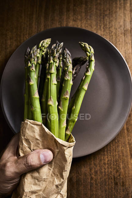 Mão segurando um pacote de espargos verdes frescos em um saco de papel — Fotografia de Stock