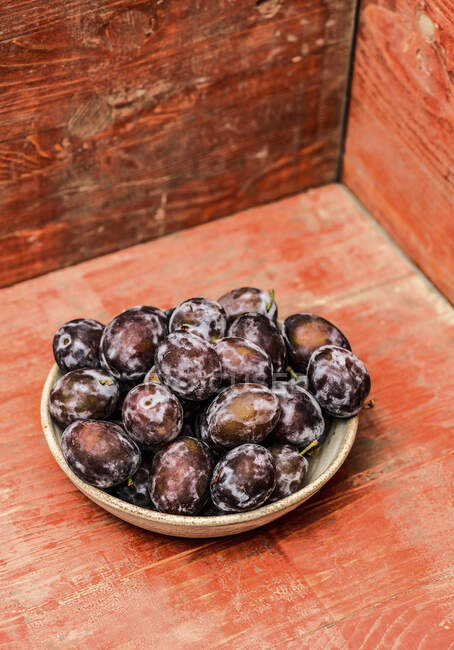 Fresh plums in bowl on red wooden surface — Stock Photo