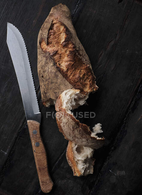 Loaf of crusty white bread and breach knife on black surface — Stock Photo