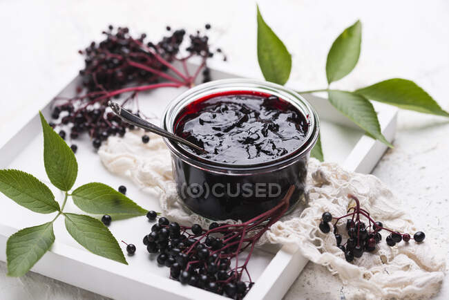 Black elderberries jam in jar with silver spoon surrounded with fresh berries and green leaves — Stock Photo