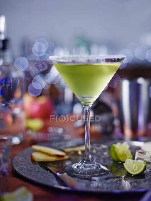 Cóctel Appletini en vaso de martini con azúcar, rodajas de manzana y cuñas de lima con cuchillo en bandeja metálica - foto de stock