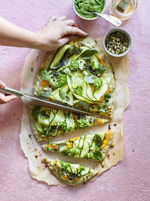 A pizza with courgetti, herbs and pesto being sliced — Stock Photo