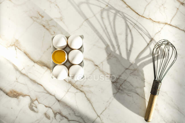Eggs in paper box, one cracked open and whisk on marble surface — Stock Photo