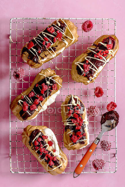 Home made eclairs with whipped cream, raspberries and chocolate on a cooling rack, view from above — Stock Photo