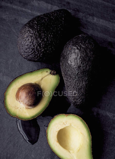Avocados on black slate, one halved — Stock Photo